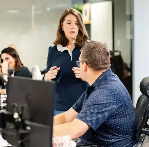 Coworker standing beside a desk speaking with a colleague seated at a computer in an office