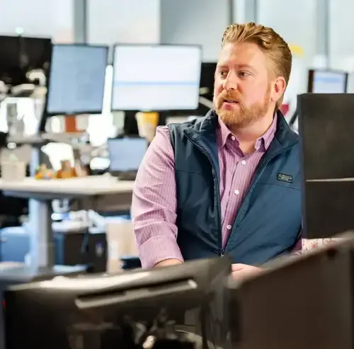 Man seated at a desk looking toward colleagues in an open office with multiple computer monitors