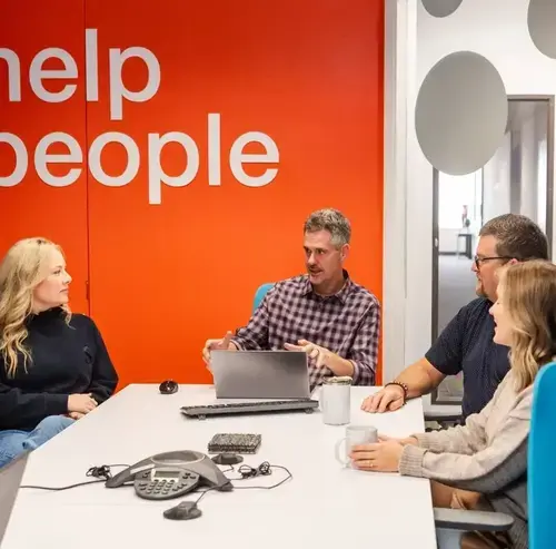 Four coworkers meeting around a conference table in an office with an orange wall reading “help people”